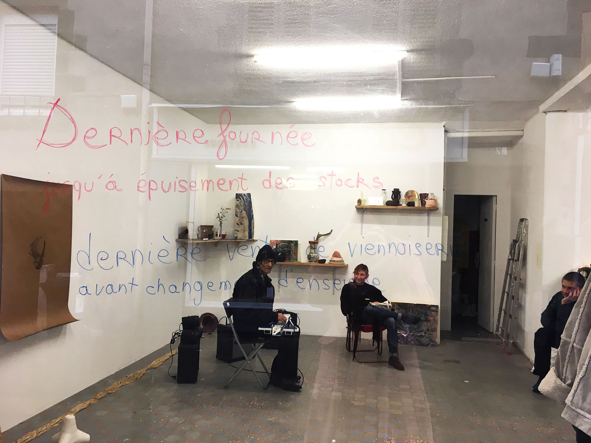 On voit des gens assis dans n espace blanc avec pleins d'œuvres d'art autour d'eux, a premier blanc un texte ecrit sur une vitre dit : Dernière fournée, jusqu'à épuisement des stocks. Dernières vente de viennoiserie avant changement d'enseigne.